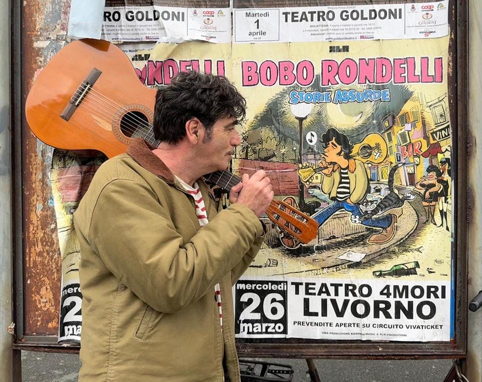 
	Il cantautore Bobo Rondelli davanti a un manifesto che pubblicizza il suo concerto a Livorno

