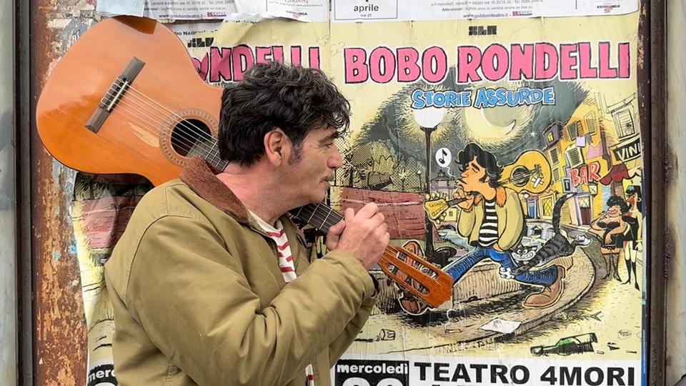 Il cantautore Bobo Rondelli davanti a un manifesto che pubblicizza il suo concerto a Livorno