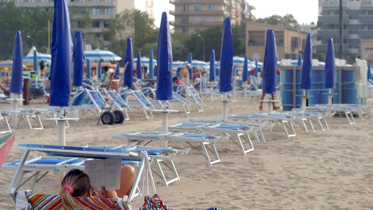 Una turista solitaria in spiaggia si legge un libro davanti ad una fila di ombrelloni chiusi
