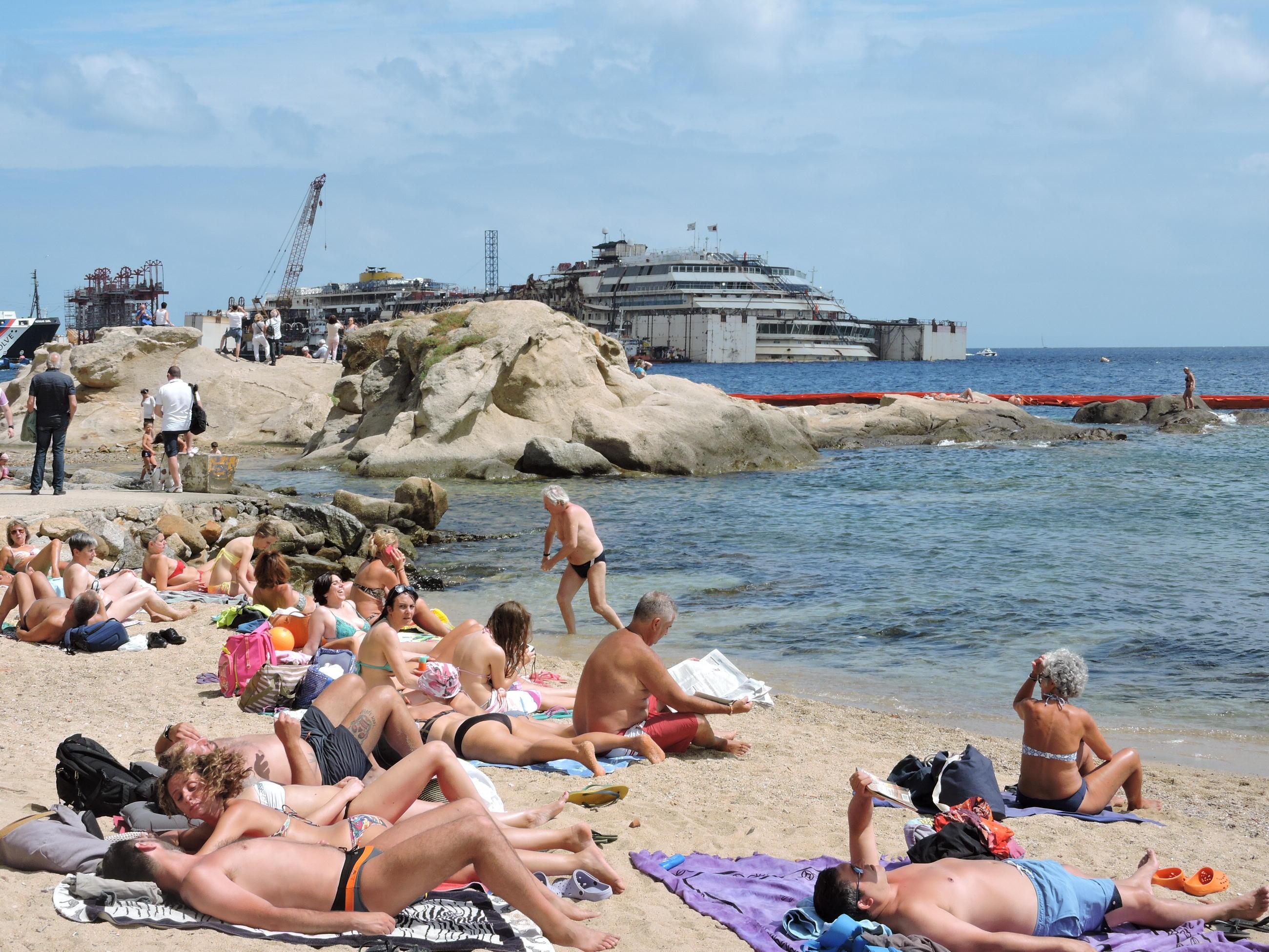 Turisti sulle spiagge di Giglio Porto con il relitto della Concordia sullo sfondo (foto Russo)