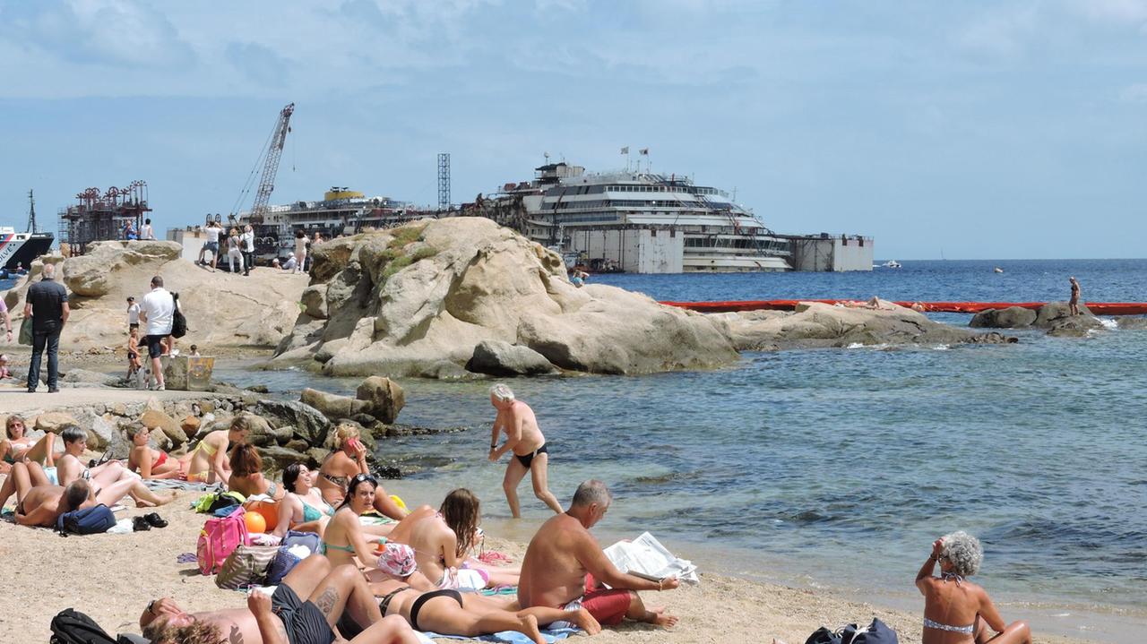 Turisti sulle spiagge di Giglio Porto con il relitto della Concordia sullo sfondo (foto Russo)