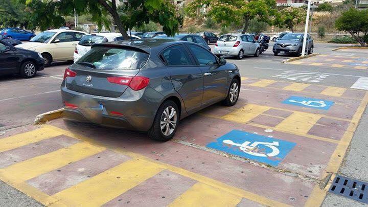 L'Alfa Romeo senza pass per disabili parcheggiata perpendicolarmente a due posteggi riservati ai portatori di handicap nel parcheggio Conad di via Jenner