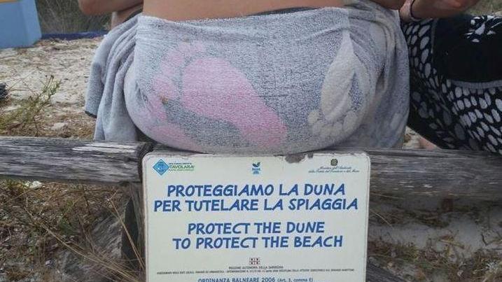 Un turista seduto sul cartello che impone il divieto di calpestare le dune