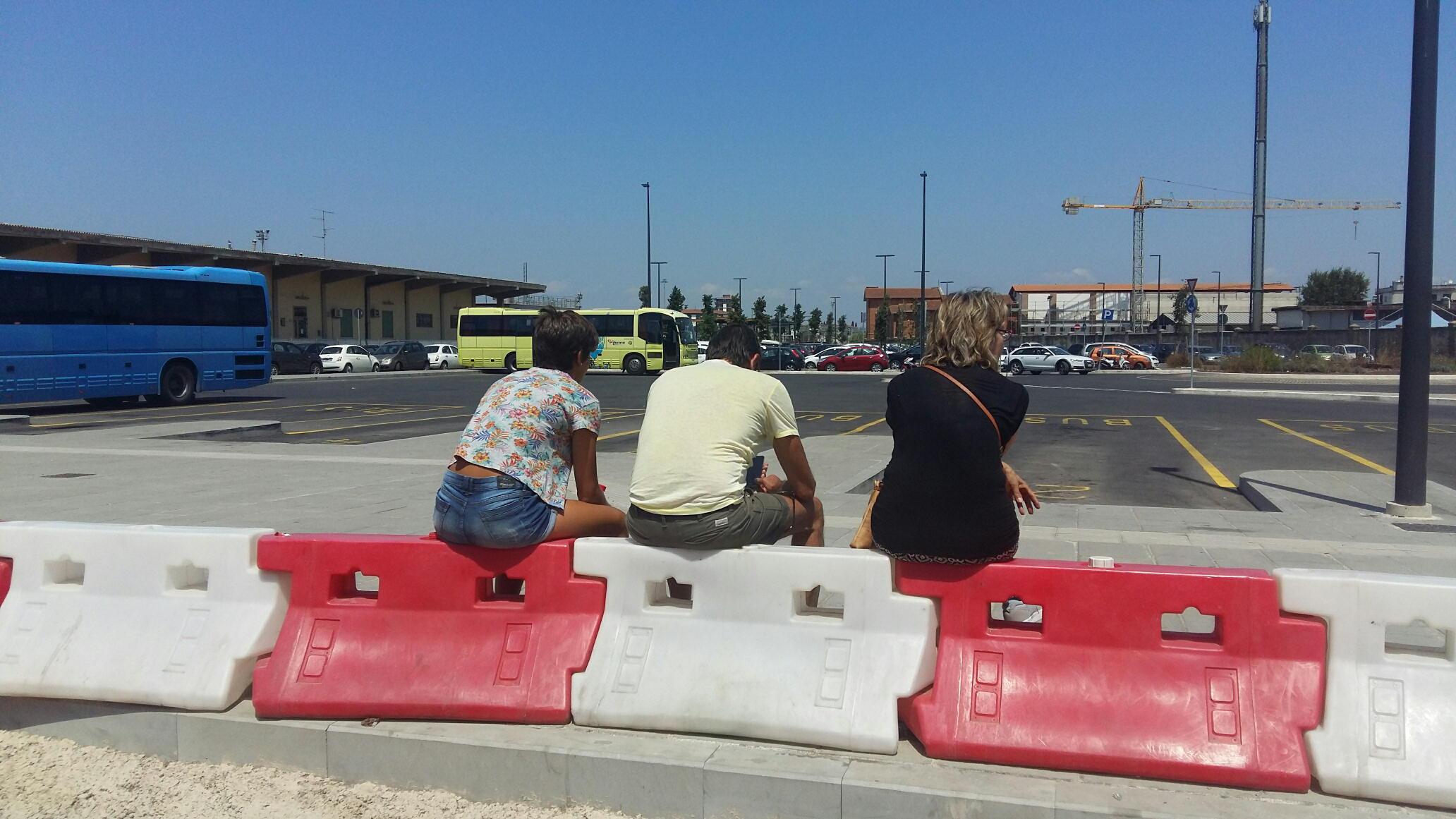 Una famiglia seduta sulle balaustre in attesa dell'autobus nel piazzale della stazione di Grosseto