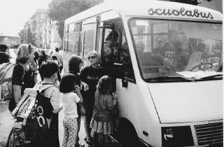 Bambini mentre salgono sul pulmino per andare a scuola e a sinistra auto di genitori fermi davanti alle scuole