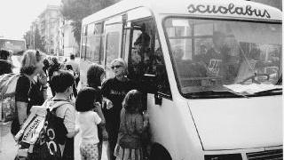 Bambini mentre salgono sul pulmino per andare a scuola e a sinistra auto di genitori fermi davanti alle scuole