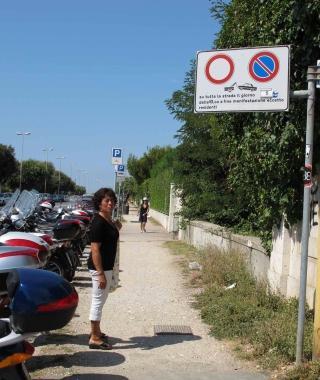 Una donna osserva stupita la segnaletica sul lungomare; decine di scooter lasciati in sosta vietata e un gruppo di bagnanti davanti ai Fiume discute del divieto  