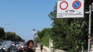 Una donna osserva stupita la segnaletica sul lungomare; decine di scooter lasciati in sosta vietata e un gruppo di bagnanti davanti ai Fiume discute del divieto
