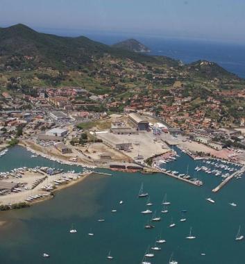 Sopra una veduta aerea dell’area portuale destinata alla realizzazione del Porto Cantieri, a sinistra il piazzale del cantiere Esaom a Portoferraio