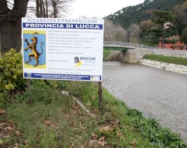 <strong>FIUME CAMAIORE.</strong> A sinistra il cartellone che annuncia i lavori sugli argini della Provincia per la messa in sicurezza; il fiume tra Ponte di Sasso e i Cavallini e a destra l’argine che verrà completamente rifatto con l’alveo raddoppiato. Sotto la strada lungofiume che sarà ridotta ad una semplice pista