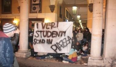 Lo striscione affisso dagli studenti al porticato di piazza Grande: dietro i ragazzi seduti in terra fanno lezione. A dx un manifesto  