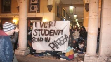 Lo striscione affisso dagli studenti al porticato di piazza Grande: dietro i ragazzi seduti in terra fanno lezione. A dx un manifesto