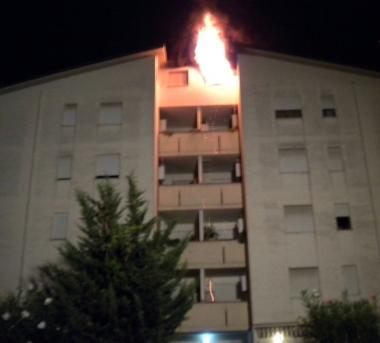 Le fiamme escono dalla mansarda A destra, due residenti “sfollati” e alle loro spalle il palazzo con l’appartamento bruciato all’ultimo piano