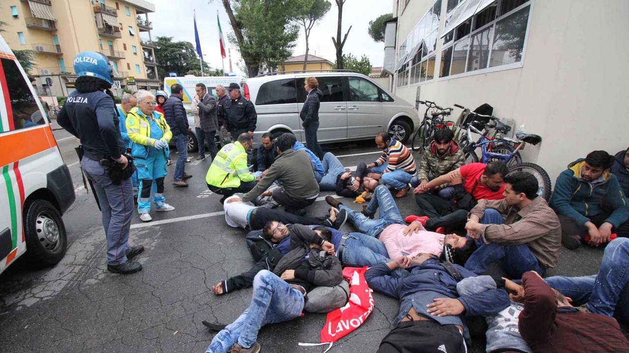 Gli operai pakistani sdraiati a terra con la polizia che li controlla e i sanitari che medicano alcuni contusi