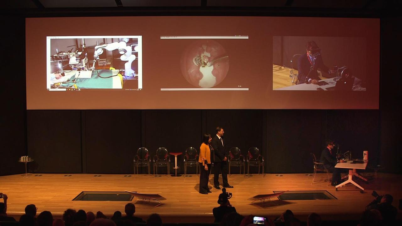Il palco del Vodafone Village: il professor Matteo Trimarchi seduto alla scrivania esegue l'intervento, il "paziente" si trova nella sala operatoria del San Raffaele