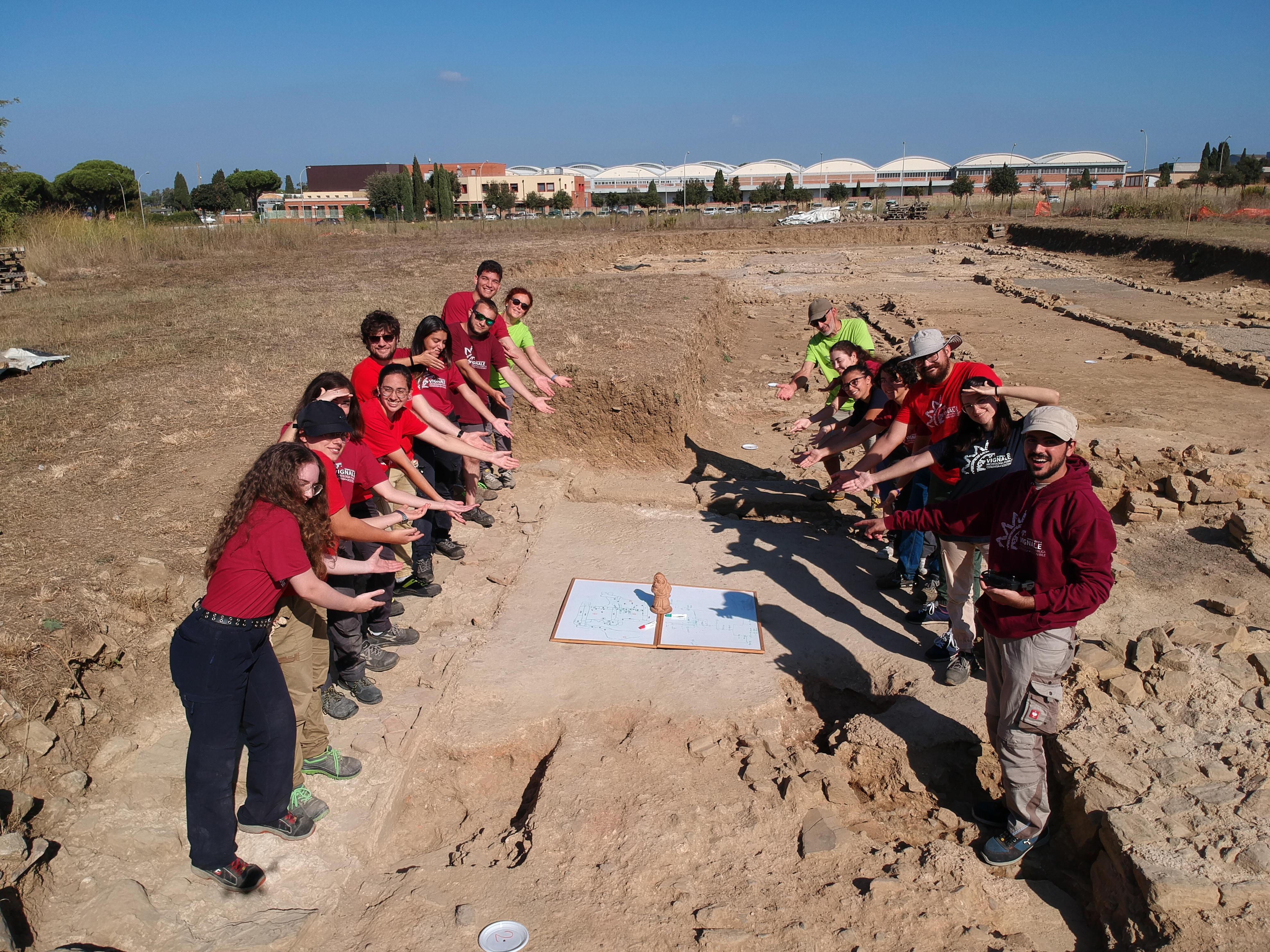 Docenti, ricercatori e studenti dell’università di Siena sul sito di Vignale indicano la lavagna su cui si stavano appuntando nuove idee sulla sequenzastratigrafica del sito, lavagna su cui poggia il nano di terracotta da tanti anni colonna portante del progetto Uomini e Cose a Vignale