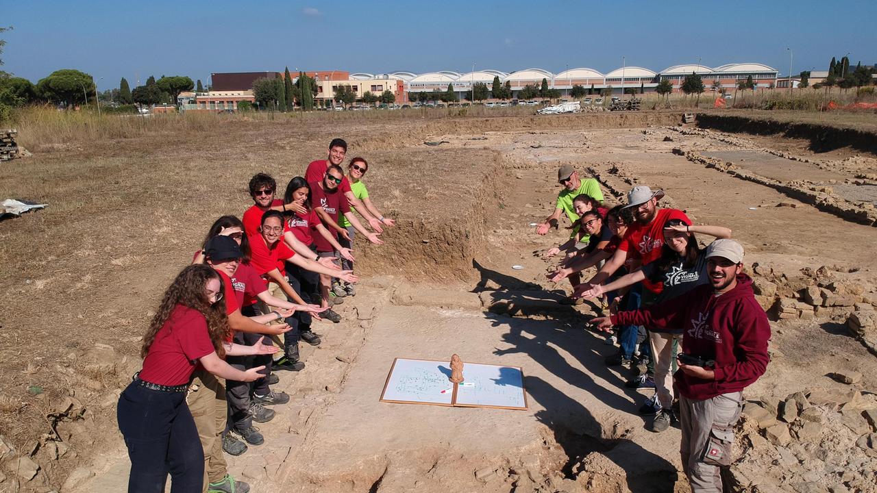 Docenti, ricercatori e studenti dell’università di Siena sul sito di Vignale indicano la lavagna su cui si stavano appuntando nuove idee sulla sequenzastratigrafica del sito, lavagna su cui poggia il nano di terracotta da tanti anni colonna portante del progetto Uomini e Cose a Vignale