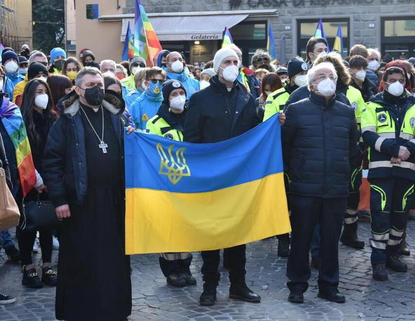 Un gruppo di persone alla manifestazione in piazza Umberto I a Castelnuovo tiene tra le mani i lembi di una bandiera ucraina, a sinistra don Lyupak, a capo dell’Esarcato